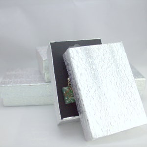 May include: A silver gift box with a textured pattern is open, revealing a black interior and a small green and white object inside.