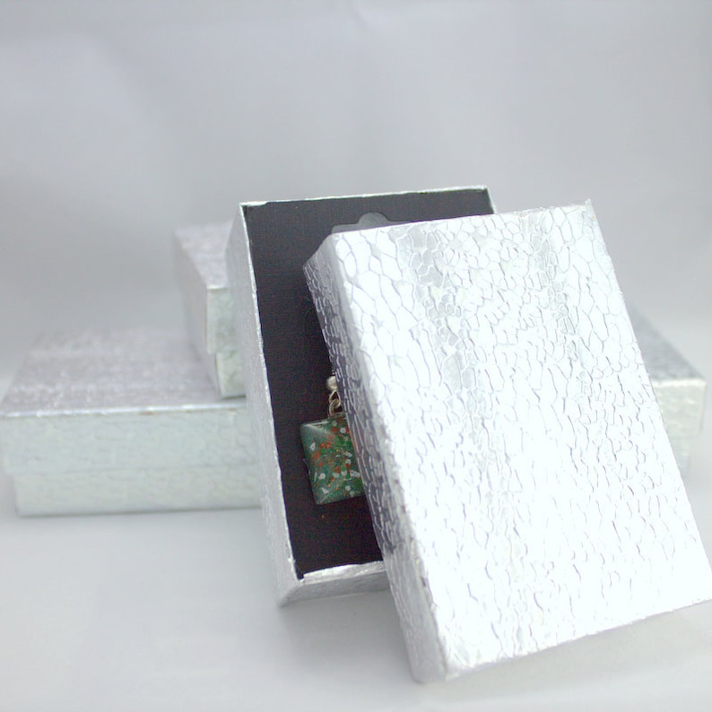 May include: A silver gift box with a textured pattern is open, revealing a black interior and a small green and white object inside.