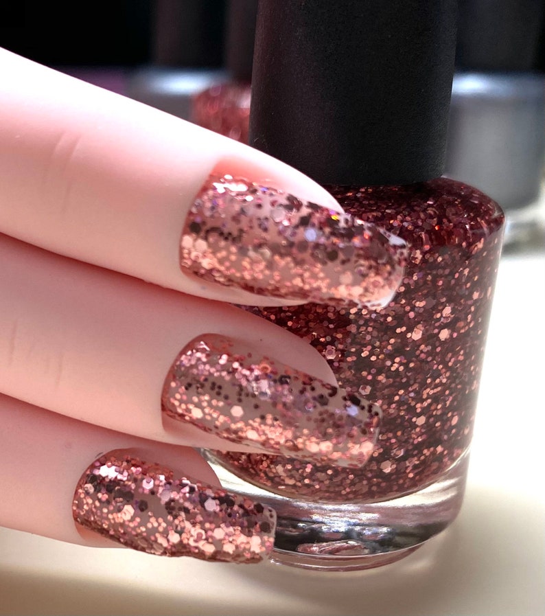 May include: A clear glass bottle of rose gold glitter nail polish with a black cap. A hand is shown with three fingernails painted with the rose gold glitter polish.