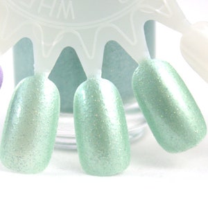 May include: A close-up of a light green nail polish with a shimmery finish. The polish is shown on a nail polish display with several other colours.