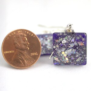 May include: Square purple glass earrings with iridescent glitter embedded in the glass. The earrings are shown next to a US penny for size comparison.