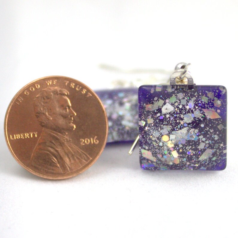 May include: Square purple glass earrings with iridescent glitter embedded in the glass. The earrings are shown next to a US penny for size comparison.