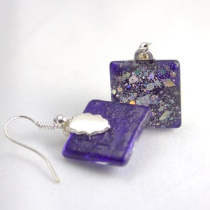 May include: A pair of square glass earrings with a purple iridescent finish and silver hooks. The earrings have a glittery texture.
