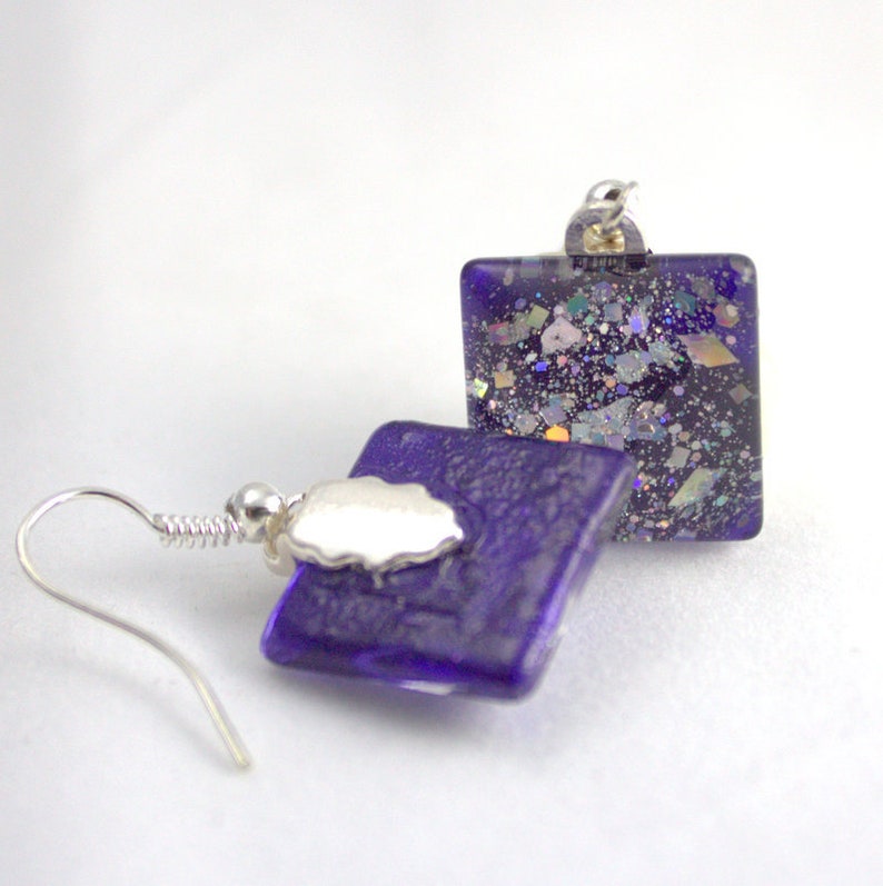 May include: A pair of square glass earrings with a purple iridescent finish and silver hooks. The earrings have a glittery texture.
