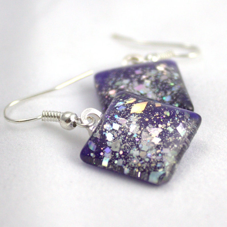 May include: A pair of square-shaped earrings with a purple glass base and iridescent glitter embedded in the glass. The earrings are attached to silver hooks.