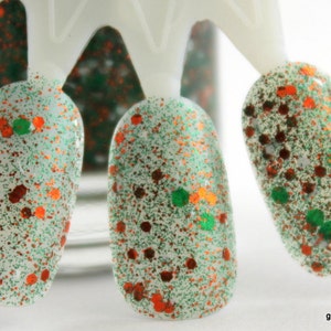 May include: Close-up of three nail polish swatches on a white display stand. The nail polish is a clear base with green and orange glitter.