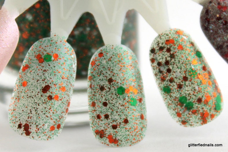 May include: Close-up of three nail polish swatches on a white display stand. The nail polish is a clear base with green and orange glitter.