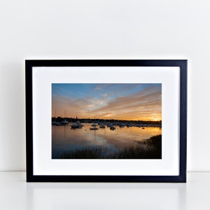 May include: A framed photograph of a sunset over a harbor with boats docked. The sky is a mix of orange, pink, and blue with clouds. The water is calm and reflects the colors of the sky.