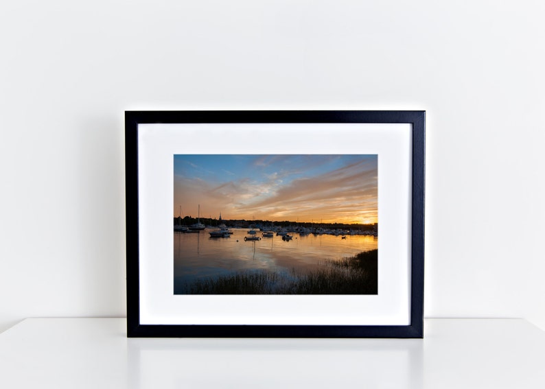 May include: A framed photograph of a sunset over a harbor with boats docked. The sky is a mix of orange, pink, and blue with clouds. The water is calm and reflects the colors of the sky.
