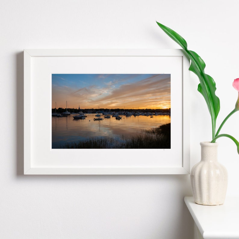 May include: A framed photograph of a sunset over a calm body of water with boats docked in the distance. The sky is a mix of orange, pink, and blue with clouds reflecting in the water.
