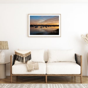 May include: A white couch with two pillows and a throw blanket in a living room with a framed photograph of a sunset over a body of water with boats.