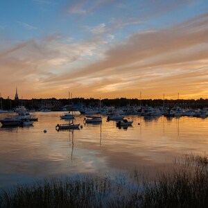 May include: A scenic view of a harbor at sunset with a variety of sailboats and motorboats docked. The sky is a vibrant orange and pink with clouds reflecting in the calm water.