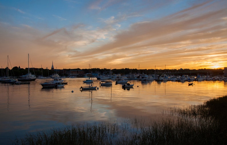 May include: A scenic view of a harbor at sunset with a variety of sailboats and motorboats docked. The sky is a vibrant orange and pink with clouds reflecting in the calm water.