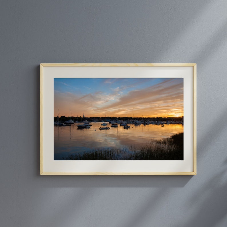 May include: A framed photograph of a sunset over a harbor with boats docked. The sky is a vibrant orange and pink, with clouds reflecting in the water.