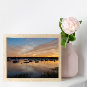 May include: A framed photograph of a harbor scene at sunset. The water is calm and reflects the orange and pink sky. There are many boats docked in the harbor.
