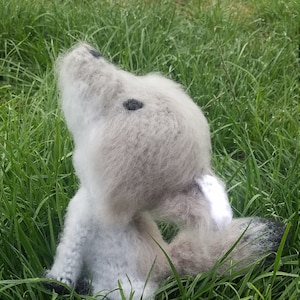 May include: A gray and white crocheted wolf plush toy sitting in green grass. The wolf has black eyes and a long tail.