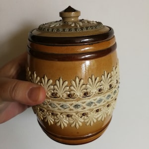 May include: A brown and white ceramic jar with a lid. The jar has a decorative design of leaves and a band of brown glaze. The lid has a knob on top.