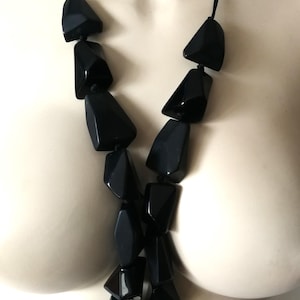 Puede incluir: Un collar negro llamativo con grandes cuentas geométricas facetadas. Las cuentas están ensartadas en un cordón negro, creando un accesorio audaz y moderno. El collar es una pieza de joyería llamativa.