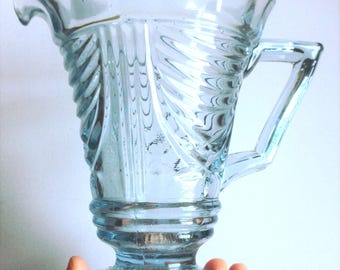 May include: A light blue glass pitcher featuring a flared rim, a sturdy angular handle, and a decorative moulded pattern of vertical ribs and swirling lines. The base is circular with a radial design, supporting the tapered body of the vessel.
