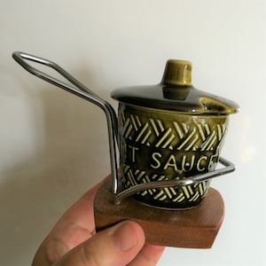 Może przedstawiać: Wazon na sos chili z zielonej ceramiki w stylu vintage z pokrywką, metalowym uchwytem i drewnianą podstawą. Pojemnik ma teksturowany wzór, a na przodzie nadrukowano napis "HOT SAUCE". Uchwyt jest wykonany ze srebrnego metalu.