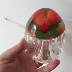 May include: A clear glass jar with a rounded shape and a matching lid featuring an embedded design of red strawberries and green leaves. The jar includes a small, clear glass spoon that rests inside, and the base has a subtle vertical ribbed texture.