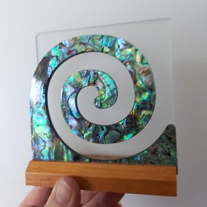 May include: A decorative spiral ornament featuring iridescent abalone shell inlay with vibrant green, blue, and purple tones. The spiral is mounted on a clear square acrylic backing and supported by a rectangular light brown wooden base stand.