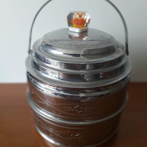 May include: A vintage metal ice bucket with a handle and lid. The bucket has a brown, textured body with silver trim and a decorative pattern. The lid is silver with a floral design, and the handle is a silver wire. The lid has a clear, square knob with a floral design.
