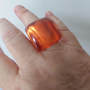 May include: A chunky, orange-amber ring with a rectangular design. The ring is made from a translucent material and is worn on a finger. It has a glossy finish and reflects light, creating a striking visual effect.