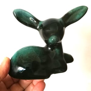 以下が含まれることがあります： 休息姿勢のシカの陶器製置物。シカは濃いティールとグリーンの色合いで釉薬がかけられており、光沢があります。大きな耳と丸みを帯びた体をしており、手で持たれています。