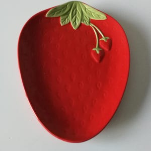 May include: A red ceramic strawberry-shaped serving dish. The dish features a textured surface and is adorned with green leaves and two small strawberries at the top. The dish is designed to resemble a strawberry.