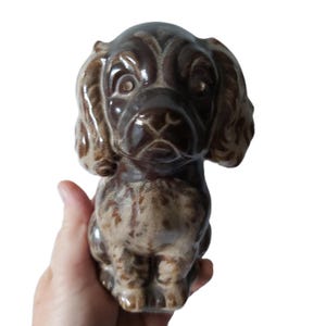May include: A ceramic dog figurine in shades of brown and tan. The dog has large, expressive eyes and floppy ears. The figurine is sitting upright, with its front paws together. A decorative item.