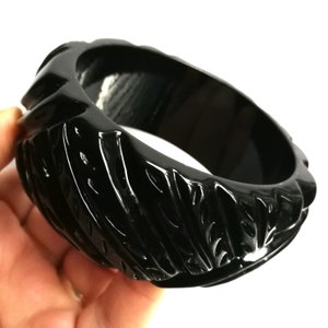 Könnte beinhalten: Ein glänzendes, schwarzes Armreif-Armband mit einem geschnitzten, strukturierten Design. Das Armband hat ein breites Band und eine glatte Innenseite. Das geschnitzte Muster erzeugt eine Reihe von Rippen und Rillen um die Außenseite des Armbands.