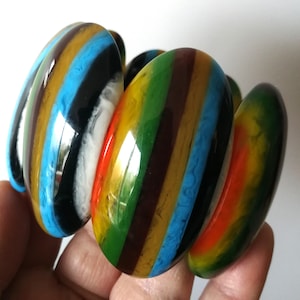 Puede incluir: Una pulsera segmentada y colorida con un acabado brillante. La pulsera presenta una variedad de colores, incluyendo rayas azules, amarillas, verdes, rojas y marrones. La pulsera está hecha de un material brillante.