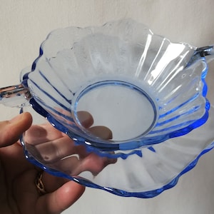 May include: A light blue glass dish and saucer set. The dish has a scalloped edge and a handle. The saucer is also scalloped. The set is made of clear glass with blue accents. The dish is approximately 4 inches in diameter.