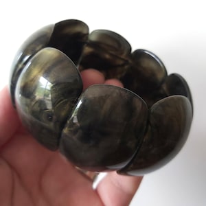 May include: A stretch bracelet composed of multiple rounded, smooth, dark charcoal-colored links with a subtle, swirling metallic gold shimmer throughout the material. The individual segments are connected to form a continuous, flexible circular band.