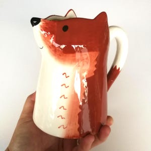 May include: A ceramic pitcher with a fox design. The pitcher is primarily white with orange and red accents, featuring a fox face on the front and a white handle. The design includes a series of red wavy lines.