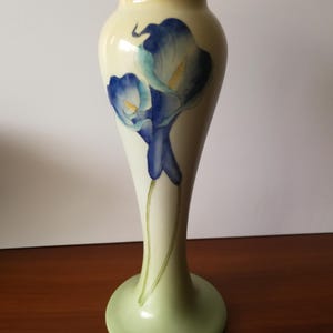 May include: A slender vase with a cream-coloured body and a flared base. It features a hand-painted design of blue calla lilies with yellow centres. The base is a soft green colour, and the vase stands on a wooden surface.
