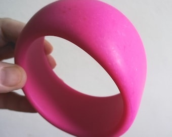 Può includere: Un braccialetto a cerchio rosa acceso. Il braccialetto liscio e arrotondato è di colore pieno e ha un design semplice e moderno. Il braccialetto è tenuto in mano su uno sfondo semplice.