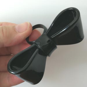May include: A glossy black plastic bow-shaped clip. The clip has a rounded design with a ring attached to the top. The clip is held by a hand against a white background.