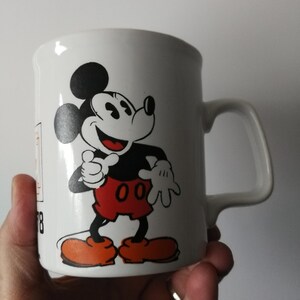 K&ouml;nnte beinhalten: Wei&szlig;er Keramikbecher mit einer Illustration von Mickey Mouse. Mickey l&auml;chelt und tr&auml;gt rote Shorts, orangefarbene Schuhe und schwarze Handschuhe. Der Becher hat einen gebogenen Griff und ein schlichtes Design.