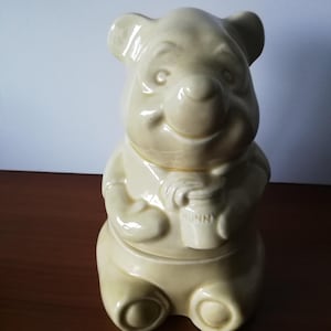 May include: A ceramic Winnie the Pooh cookie jar in a light yellow color. The jar is shaped like the character, holding a honey pot with the word "Hunny" on it. The jar has a glossy finish and is sitting on a brown surface.