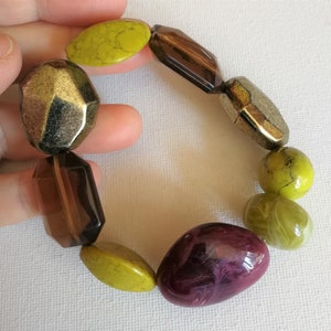 May include: A chunky bracelet featuring a variety of large beads in shades of gold, brown, green, and purple. The beads have different shapes, including faceted, oval, and round. The bracelet is held by a hand.