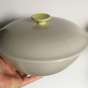 Puede incluir: Un tazón grande de servir de cerámica gris claro con una tapa a juego. La tapa tiene un pequeño pomo amarillo brillante en la parte superior.