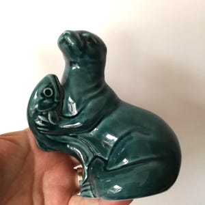 Puede incluir: Escultura de cerámica de color verde azulado de una foca sosteniendo un pez. La foca está representada con la cabeza levantada y el pez se sujeta cerca de su cuerpo. La escultura tiene un acabado brillante, sobre fondo blanco.