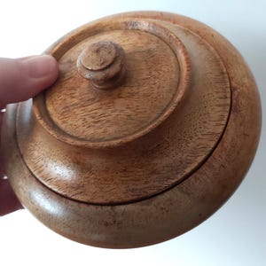 May include: A round wooden box with a lid. The box and lid are made of light brown wood. The lid has a small knob on top for easy opening. The box is held by a hand.