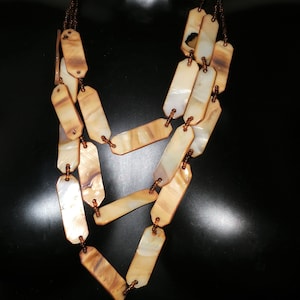 May include: A layered necklace with rectangular, iridescent shell pendants in cream and brown hues. The pendants are linked by small, brown beads and fine chains. The necklace is shown against a dark backdrop.