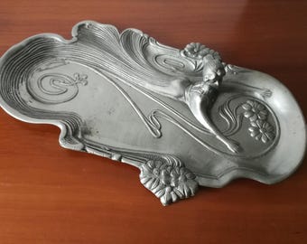 Könnte beinhalten: Eine silberfarbene Art-Nouveau-Schale mit einer skulptierten weiblichen Figur und floralen Akzenten. Die Schale hat eine längliche, asymmetrische Form mit fließenden Linien und detaillierten Reliefarbeiten. Die Schale ist aus Metall.