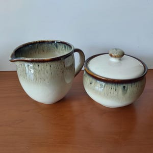 May include: A ceramic creamer jug and a matching lidded sugar bowl. Both pieces feature a cream-coloured base that transitions into a dark brown rim, with a decorative drip glaze effect in shades of brown and grey cascading down the sides.