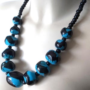 Op de afbeelding: Een opvallende ketting met grote, glanzende kralen in zwart en turkooisblauw. De kralen zijn onregelmatig van vorm en geregen op een zwart koord. Een opvallend accessoire, perfect om elke outfit op te fleuren.