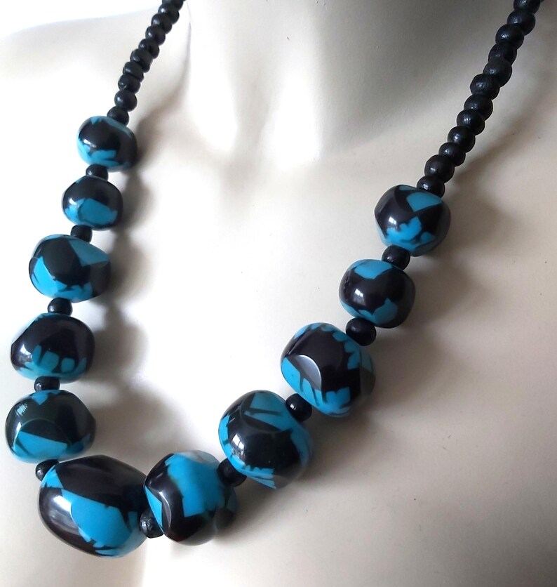 Op de afbeelding: Een opvallende ketting met grote, glanzende kralen in zwart en turkooisblauw. De kralen zijn onregelmatig van vorm en geregen op een zwart koord. Een opvallend accessoire, perfect om elke outfit op te fleuren.